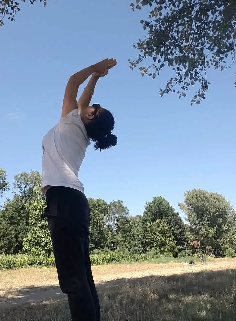 Nunzia in un piegamento all'indietro su un prato, con gli alberi e un cielo azzurro limpido alle spalle